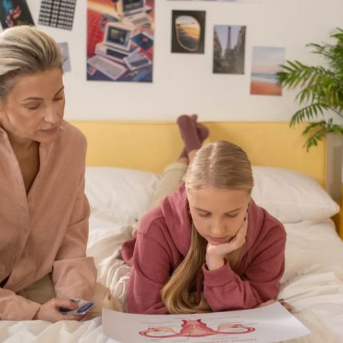 A mother and daughter talk over illustrated materials in a cozy bedroom setting.