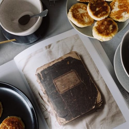 Top view of plate with delicious curd pancakes placed on recipe book surrounded by various plates on gray tabletop in kitchen
