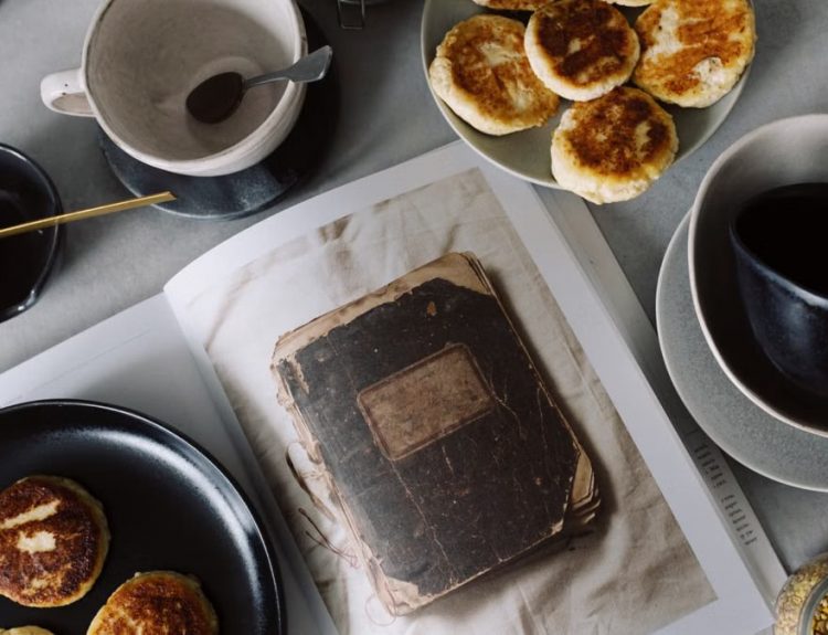 Top view of plate with delicious curd pancakes placed on recipe book surrounded by various plates on gray tabletop in kitchen