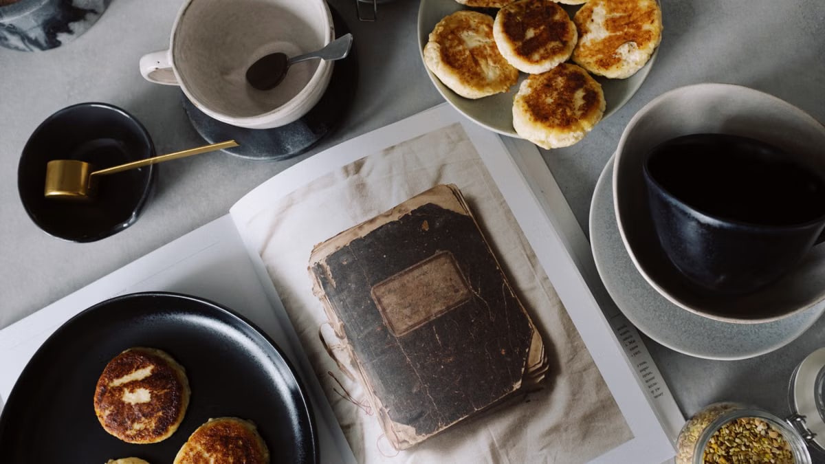 Top view of plate with delicious curd pancakes placed on recipe book surrounded by various plates on gray tabletop in kitchen