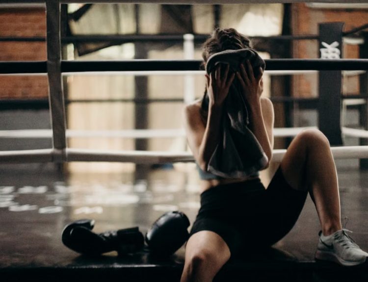 A female boxer sitting in a gym ring, resting and wiping sweat with a towel after a workout.